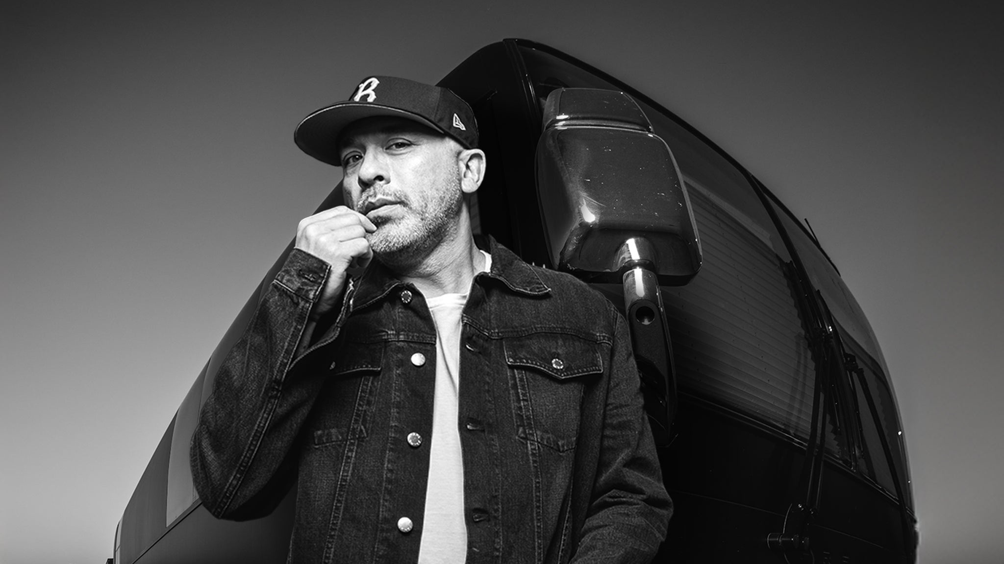 Schwarz-Weiß-Foto von Jo Koy in dunkler Kappe und Jacke neben einem Fahrzeug, eine Hand nahe am Gesicht.