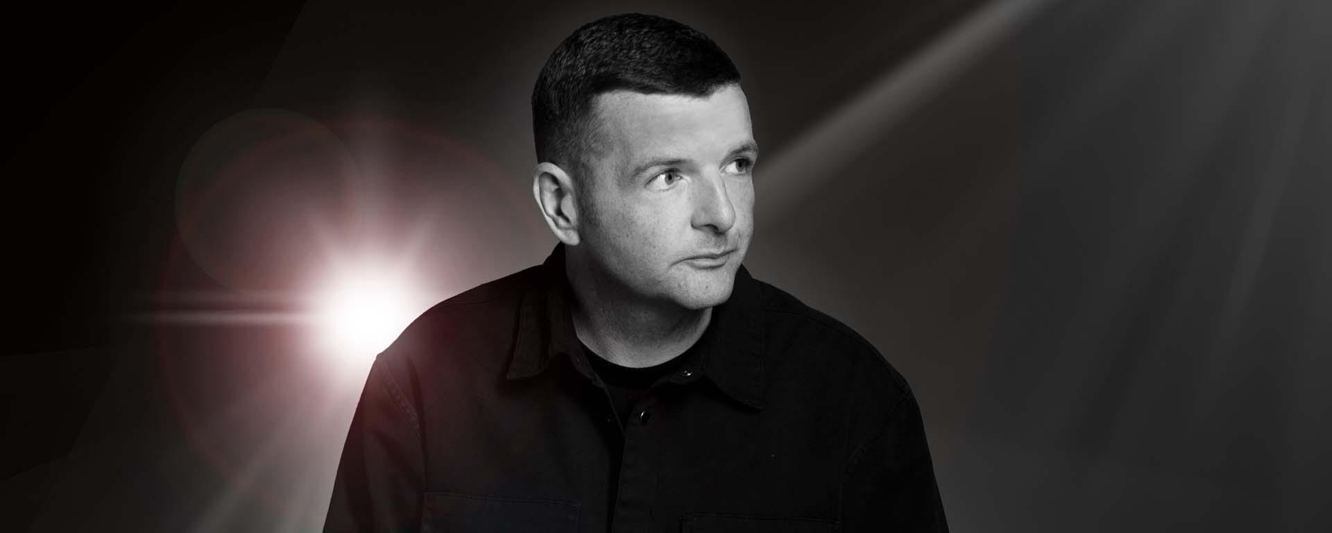 Schwarz-weißes Werbeporträt von Kevin Bridges im dunklen Oberteil, seitlich blickend, mit hellem Lichtreflex vor dunklem Hintergrund.